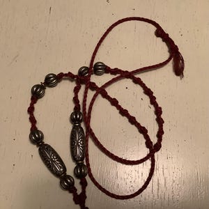 May include: A burgundy necklace featuring silver-coloured metal beads. The beads are in various shapes, including ribbed spheres and elongated, patterned ovals. The necklace is made of a twisted cord.