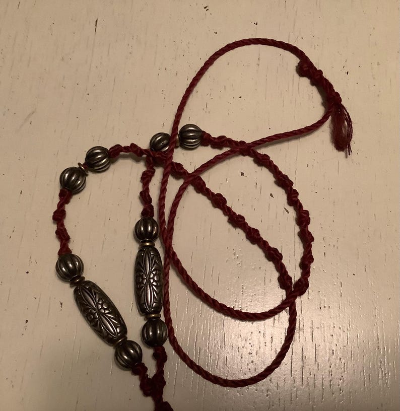 May include: A burgundy necklace featuring silver-coloured metal beads. The beads are in various shapes, including ribbed spheres and elongated, patterned ovals. The necklace is made of a twisted cord.