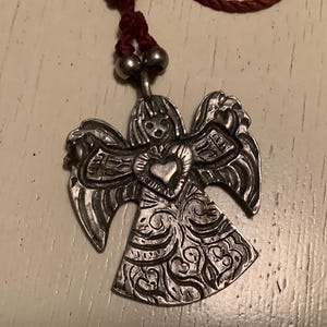 May include: Silver angel pendant with detailed wings and dress, featuring a heart-shaped centre. The pendant hangs from a maroon cord necklace with silver beads.