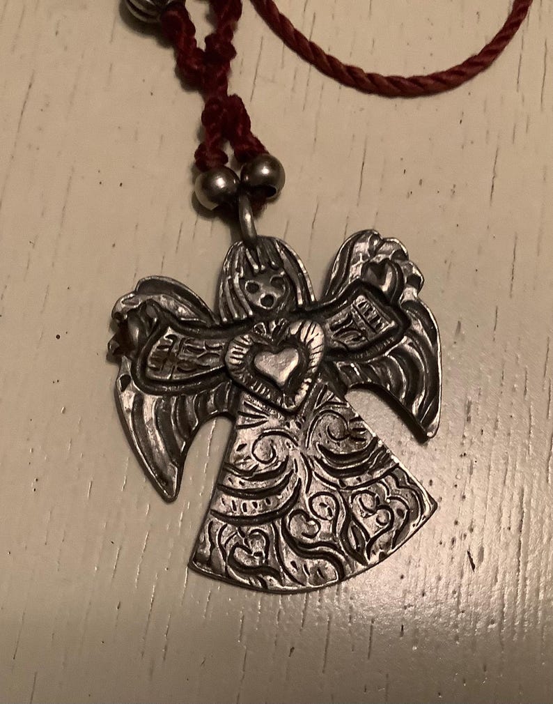 May include: Silver angel pendant with detailed wings and dress, featuring a heart-shaped centre. The pendant hangs from a maroon cord necklace with silver beads.