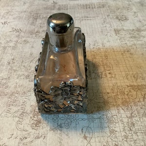 May include: A clear glass bottle with a silver top and a silver decorative frame. The frame has a floral design.