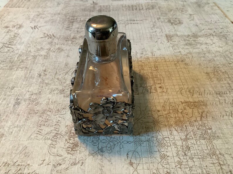 May include: A clear glass bottle with a silver top and a silver decorative frame. The frame has a floral design.