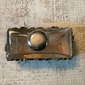 May include: A silver-toned glass bottle stopper with a decorative, ornate design. The stopper has a round, silver-toned knob on top.