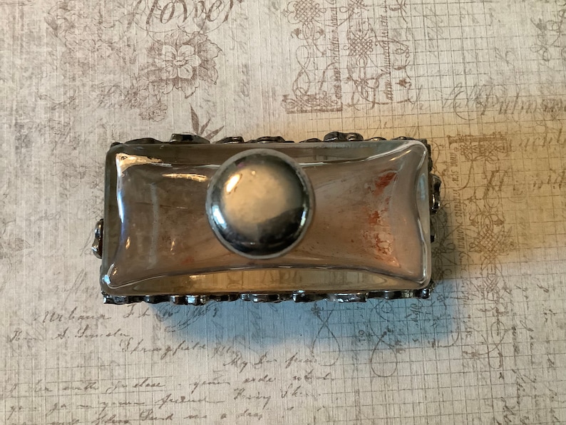 May include: A silver-toned glass bottle stopper with a decorative, ornate design. The stopper has a round, silver-toned knob on top.