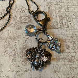 May include: A bronze chain necklace with a bee charm, a crystal pendant, and a crystal charm. The necklace is on a white background with a grid pattern.