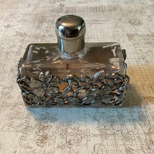 May include: A clear glass bottle with a silver top and a silver floral design around the base. The bottle is filled with a clear liquid.