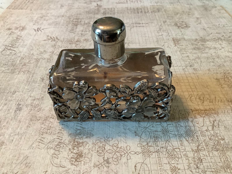 May include: A clear glass bottle with a silver top and a silver floral design around the base. The bottle is filled with a clear liquid.