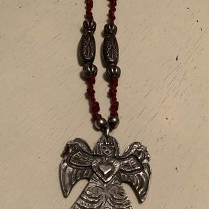 May include: A silver angel pendant necklace with a red cord. The angel pendant has detailed wings and a heart-shaped centre. The necklace features decorative silver beads.