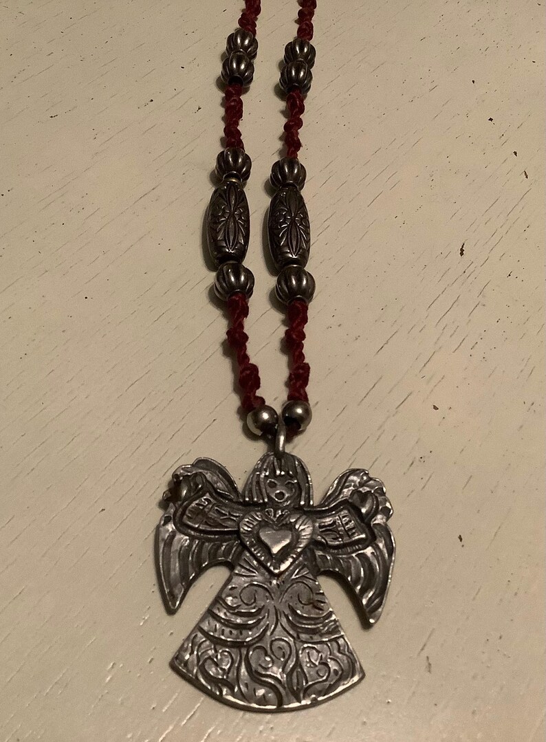 May include: A silver angel pendant necklace with a red cord. The angel pendant has detailed wings and a heart-shaped centre. The necklace features decorative silver beads.