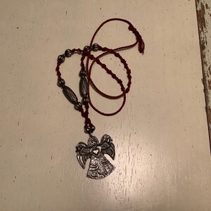 May include: A silver angel pendant necklace with a burgundy cord. The necklace features decorative beads and a detailed angel pendant. The angel has outstretched wings and intricate patterns. The necklace is displayed on a white surface.