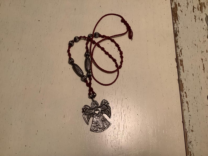 May include: A silver angel pendant necklace with a burgundy cord. The necklace features decorative beads and a detailed angel pendant. The angel has outstretched wings and intricate patterns. The necklace is displayed on a white surface.