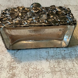 May include: A clear glass bottle with a silver metal floral design around the top and bottom. The bottle is sitting on a patterned surface.