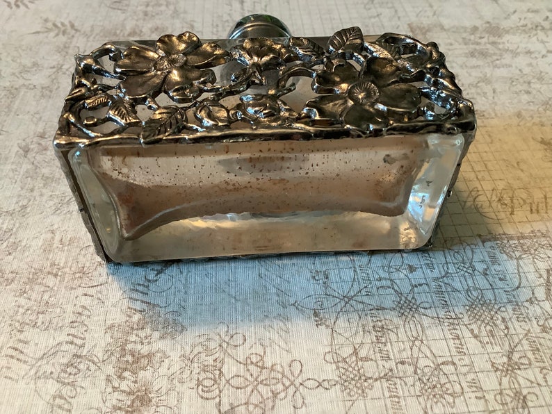 May include: A clear glass bottle with a silver metal floral design around the top and bottom. The bottle is sitting on a patterned surface.