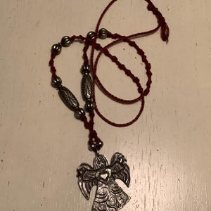 May include: A silver angel pendant necklace with a red cord. The pendant features an angel with detailed wings and a heart in the centre. The necklace includes silver beads and a knotted cord.