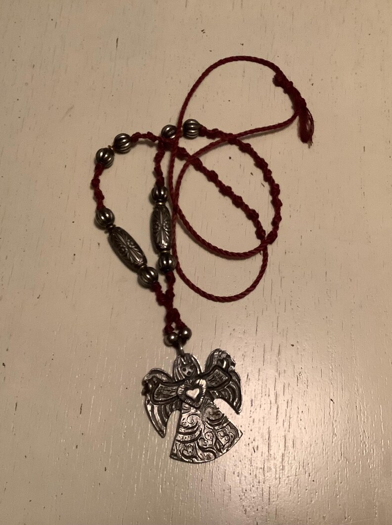 May include: A silver angel pendant necklace with a red cord. The pendant features an angel with detailed wings and a heart in the centre. The necklace includes silver beads and a knotted cord.