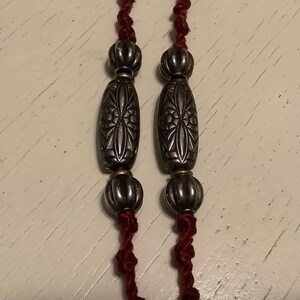 May include: Close-up of a necklace featuring silver-toned beads and decorative oval-shaped pendants. The beads are strung on a dark red cord. The pendants have an etched floral design. The necklace is displayed against a light-coloured surface.