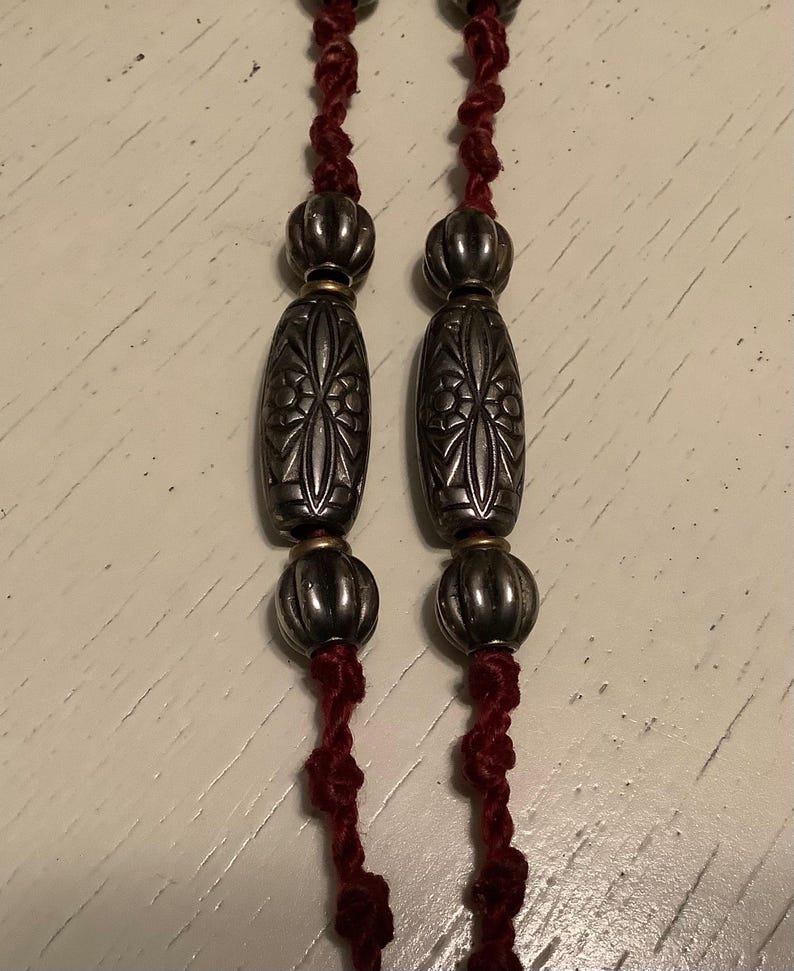 May include: Close-up of a necklace featuring silver-toned beads and decorative oval-shaped pendants. The beads are strung on a dark red cord. The pendants have an etched floral design. The necklace is displayed against a light-coloured surface.