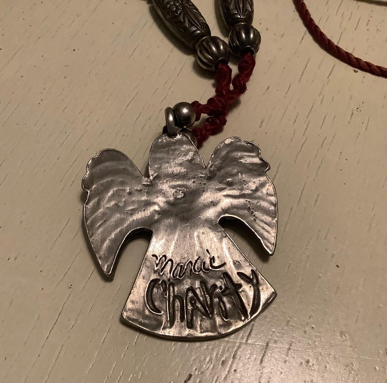 May include: A silver angel pendant necklace with the words "Marie Charity" inscribed on the pendant. The necklace has a red cord and silver beads.