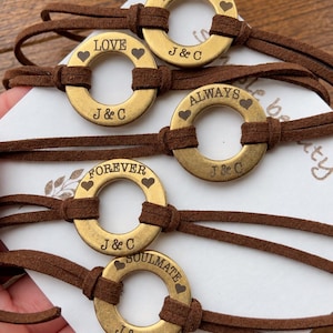 Puede incluir: Cinco pulseras de cordón de ante marrón con dijes circulares de metal en tono latón. Cada dije está grabado con las iniciales J & C y una palabra única: TOGETHER, LOVE, ALWAYS, FOREVER o SOULMATE, junto a pequeños corazones.