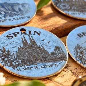 May include: A circular silver-toned metal charm featuring an engraved illustration of the Sagrada Familia in Spain, with the text SPAIN and the coordinates 41.3851 N, 2.1734 E etched below, accompanied by floral accents and two small birds.