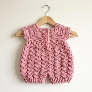 Puede incluir: Un pelele de ganchillo rosa empolvado con un patrón de punto de concha texturizado en el cuerpo, un canesú sólido y tres botones pequeños en la parte central trasera. La prenda se muestra en una percha de madera clara sobre un fondo blanco roto.