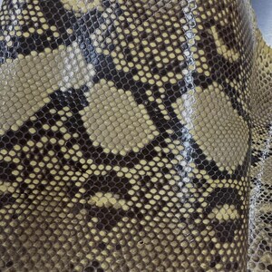 May include: A sheet of textured snakeskin leather featuring a repeating pattern of dark brown and tan scales. The surface has a glossy finish with a distinct, organic reptilian scale texture that covers the entire visible area of the material.