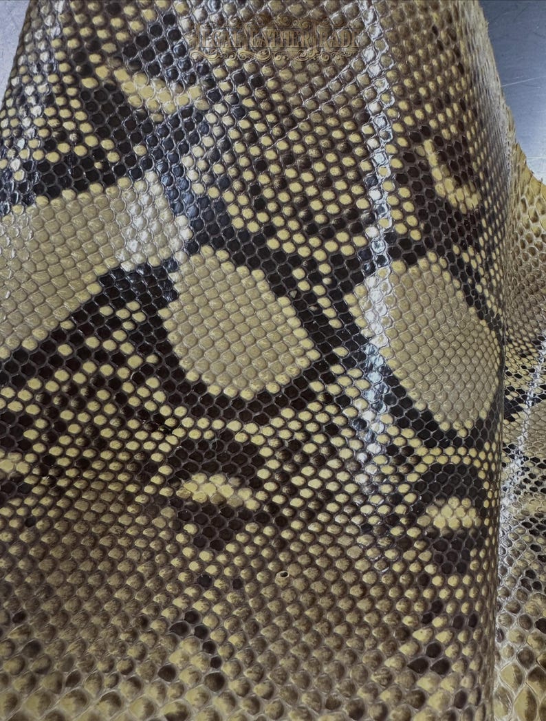 May include: A sheet of textured snakeskin leather featuring a repeating pattern of dark brown and tan scales. The surface has a glossy finish with a distinct, organic reptilian scale texture that covers the entire visible area of the material.