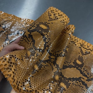May include: A flexible sheet of genuine snakeskin leather featuring a vibrant mustard yellow base color with a repeating pattern of dark brown and black diamond-shaped scales. The textured surface displays the natural scale structure of the hide.