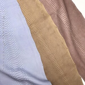 May include: Three strips of textured snake skin leather in light blue, tan, and dusty rose. The material features a repeating scale pattern throughout. The text "Zaragoza Tannery" is printed vertically along the left edge of the blue leather.