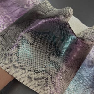Puede incluir: Un trozo de piel de serpiente con un patrón de escamas texturizado en un degradado de tonos grises, blancos y morados iridiscentes. El material muestra un brillo reflectante en la superficie. El texto LEGAL LEATHER TRADE es visible arriba.