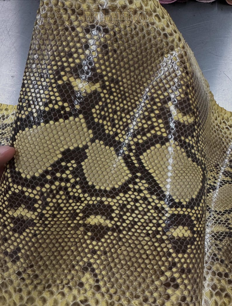 May include: A piece of natural snakeskin leather featuring a repeating diamond-shaped scale pattern in shades of yellow, tan, and dark brown. The surface has a glossy finish and displays the text "LEGAL LEATHER FADE" printed near the top edge.