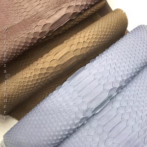May include: Three stacked pieces of textured snakeskin leather in shades of dusty rose, tan, and light blue. The material features a distinct overlapping scale pattern. The text Zaragoza Tannery is printed vertically along the left side.