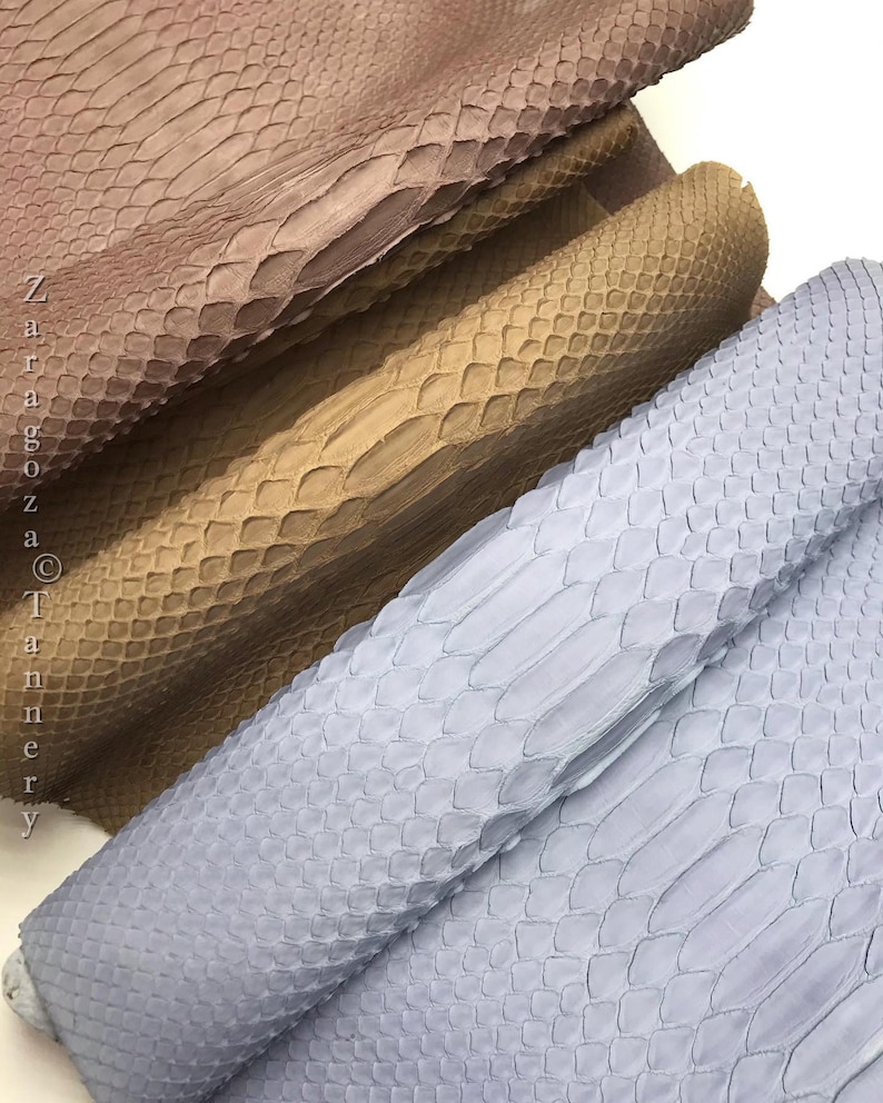 May include: Three stacked pieces of textured snakeskin leather in shades of dusty rose, tan, and light blue. The material features a distinct overlapping scale pattern. The text Zaragoza Tannery is printed vertically along the left side.