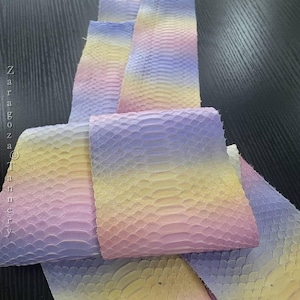 May include: A strip of genuine snakeskin leather featuring a gradient color pattern that transitions from soft pastel purple to yellow and light pink. The material displays a distinct, overlapping scale texture throughout the surface.