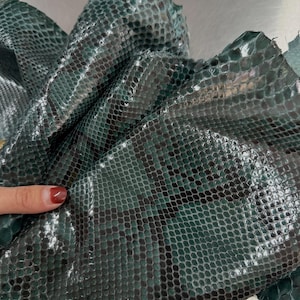 Puede incluir: Un trozo de piel de serpiente de color verde azulado oscuro y negro con un acabado brillante y un patrón de escamas distintivo. El material está doblado, mostrando su textura y flexibilidad. El texto "LEGAL LEATHER TRADE" es visible.