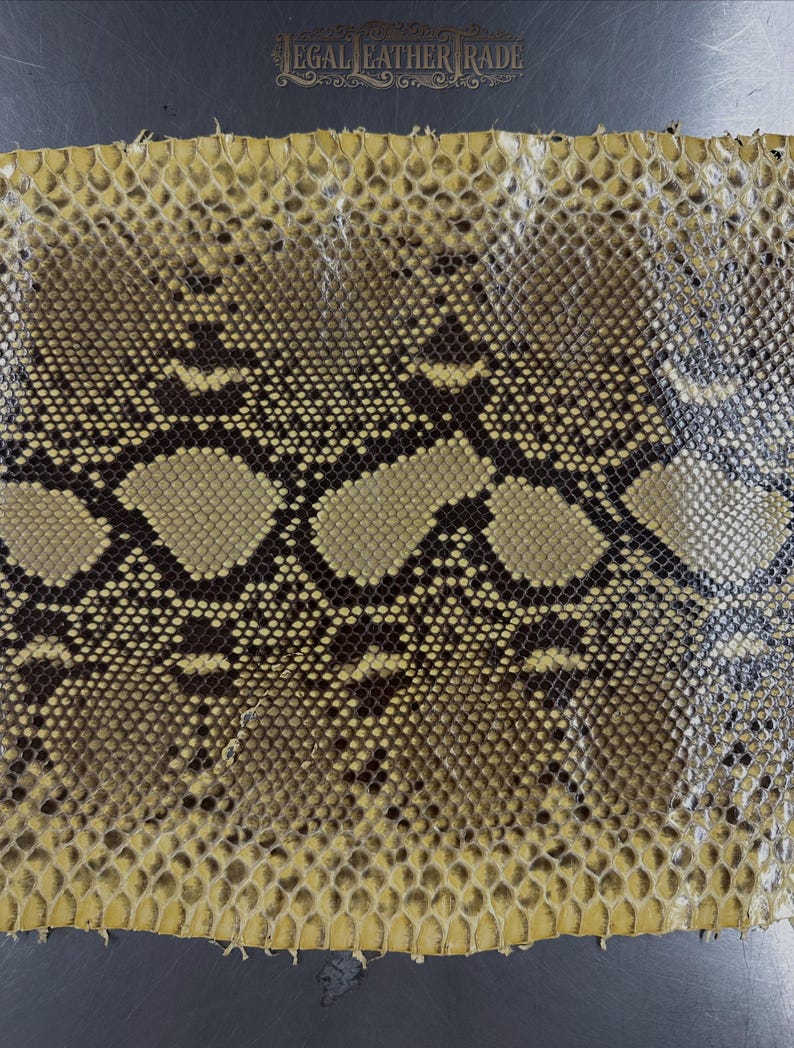 May include: A rectangular piece of snakeskin leather featuring a repeating diamond pattern in shades of tan, brown, and black. The surface has a textured, scaled appearance. The text "LEGAL LEATHER TRADE" is printed at the top center.