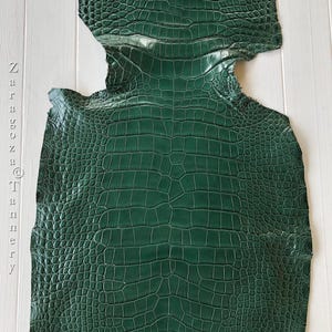 May include: A dark green crocodile skin leather hide with a glossy finish and a distinct scale pattern. The text Zaragoza Tannery is printed vertically in a light gray font along the left side of the leather piece.