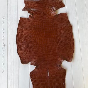 May include: A brown crocodile skin hide featuring a distinct, raised scale pattern across the entire surface. The leather has a glossy finish and is oriented vertically. The text Zaragoza Tannery is printed vertically on the left side.