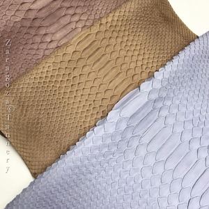 Puede incluir: Tres piezas rectangulares de cuero texturizado con un patrón de piel de serpiente en tonos malva, bronceado y azul claro. El texto "Zaragoza Tannery" está impreso verticalmente a lo largo del lado izquierdo de la pieza malva en una fuente negra simple.