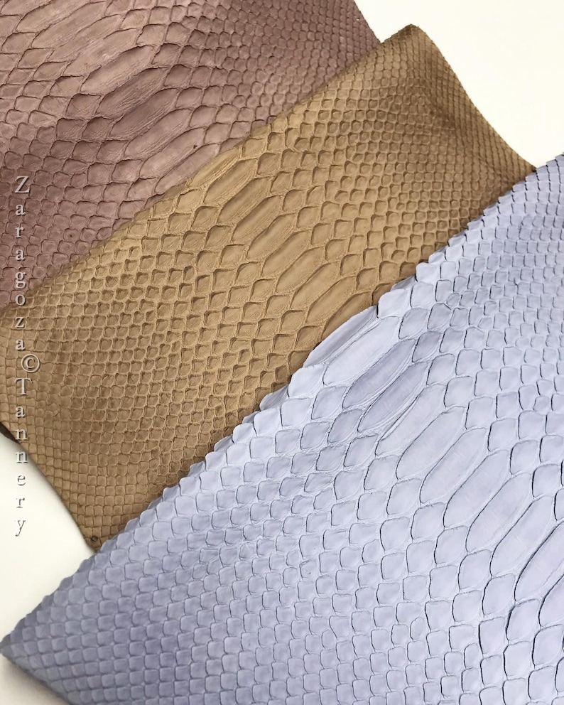 May include: Three rectangular pieces of textured leather with a snakeskin pattern in shades of mauve, tan, and light blue. The text "Zaragoza Tannery" is printed vertically along the left side of the mauve piece in a simple black font.