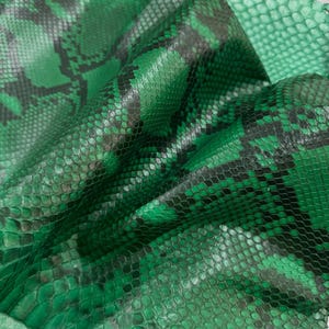 Puede incluir: Un trozo de piel de serpiente de color verde esmeralda con un patrón de escamas negras. El material tiene una superficie texturizada de escamas superpuestas e incluye una marca de agua en la parte superior central que dice LEGAL LEATHER TRADE.
