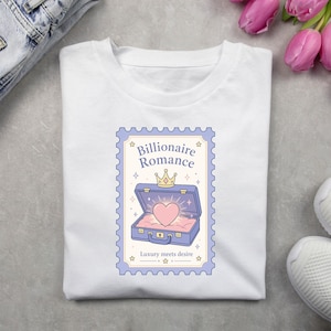 May include: A white t-shirt featuring a graphic design shaped like a postage stamp. The design shows an open purple suitcase containing a glowing pink heart and a gold crown, with the text Billionaire Romance and Luxury meets desire.
