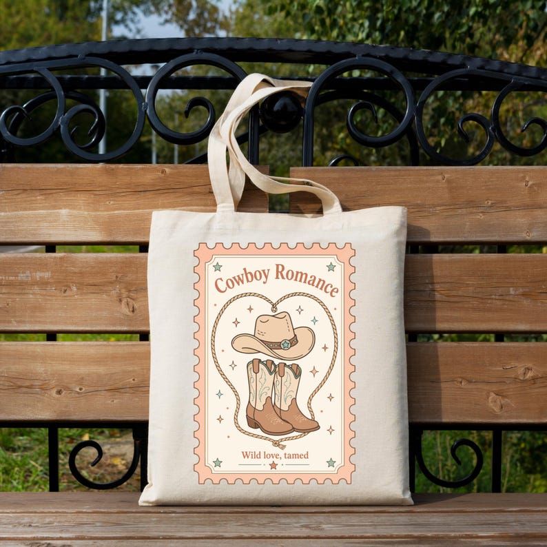 May include: A beige canvas tote bag featuring a rectangular stamp-style graphic. The design includes a cowboy hat and a pair of boots centered within a rope heart, with the text Cowboy Romance and Wild love, tamed printed in brown ink.