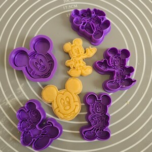 Può includere: Una collezione di cinque tagliabiscotti in plastica viola e due sagome di pasta gialla a forma di Mickey Mouse. Gli stampini mostrano varie pose del personaggio, inclusi ritratti e figure a corpo intero, disposti su un tappetino grigio.