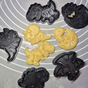Peut inclure: Une collection d'emporte-pièces en plastique noir en forme de dinosaures et des découpes de pâte jaune pâle assorties. L'ensemble comprend diverses espèces de dinosaures et une forme circulaire avec un motif de dinosaure sur un tapis en silicone.
