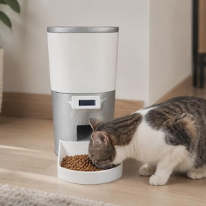 May include: A cylindrical automatic pet feeder with a white upper storage container and a silver base. The device features a small rectangular digital display screen on the front and a white feeding bowl at the bottom filled with brown dry kibble.