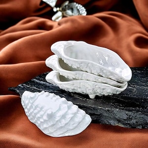 Può includere: Un set di piatti in ceramica bianca a forma di conchiglie di ostrica. I piatti presentano un esterno strutturato e realistico che imita la superficie ruvida di una conchiglia naturale e un interno liscio e lucido. Un piatto è posto separatamente.