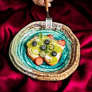 Può includere: Un piatto rotondo in ceramica artigianale con un bordo marrone testurizzato e maculato e un centro turchese lucido e crepato. Il piatto contiene una fetta di pane tostato guarnita con crema bianca, uva verde, mirtilli e fragole.