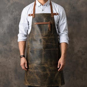 Könnte beinhalten: Eine dunkelbraune Lederschürze in Distressed-Optik mit verstellbaren, hellbraunen Lederriemen, die mit Metallnieten befestigt sind. Die Schürze verfügt über eine kleine Brusttasche mit Riemenverschluss sowie eine große aufgesetzte Tasche.