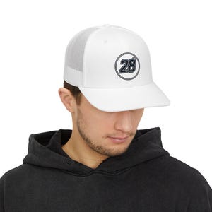 Puede incluir: Una gorra tipo trucker blanca con parte trasera de malla y panel frontal sólido. En el centro, un gráfico circular muestra el número 28 en azul oscuro con un borde fino, coronado por un pequeño icono de corona dentro del círculo.